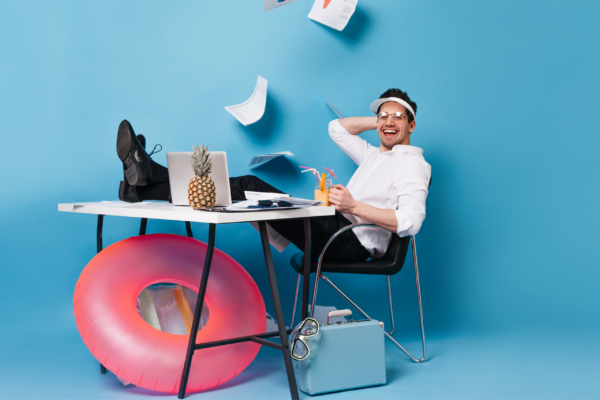 cheerful man with a white shirt and black pants enjoying cocktail on blue background with an inflatable and suitcase at his desk.
